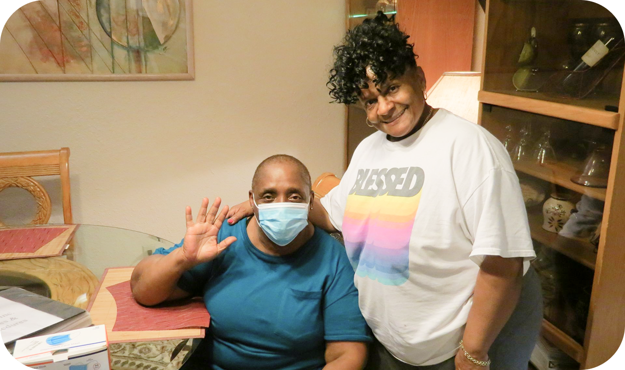A man sitting down, wearing a mask waving hi as a woman standing next to him